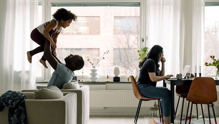 Pappa lyfter barnet upp med armarna och mamma sitter och jobbar vid en dator.