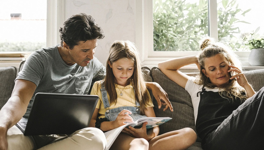 Barnfamilj som sitter och g&ouml;ra olika saker i en soffa hemma.